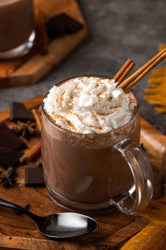 A mug of pumpkin spice hot chocolate topped with whipped cream and garnished with cinnamon sticks.