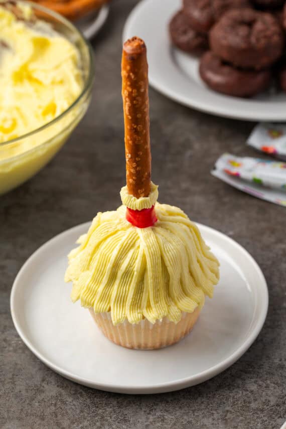 A Halloween cupcake decorated as a witch's broom on a white plate, with a bowl of yellow frosting and a plate of chocolate mini donuts in the background.