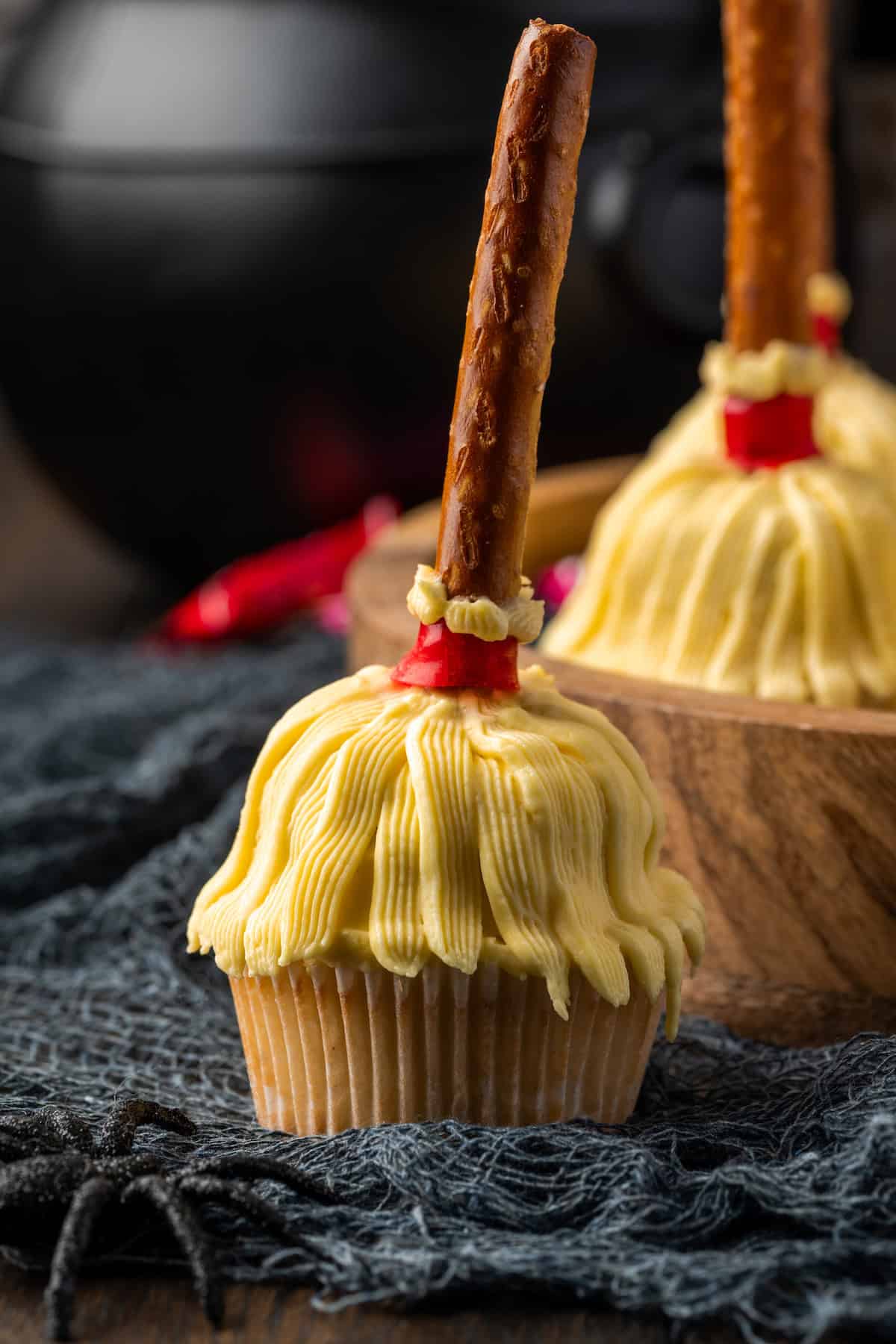 Side view of a witch's broom cupcake with more decorated Halloween cupcakes on a wooden tray in the background,