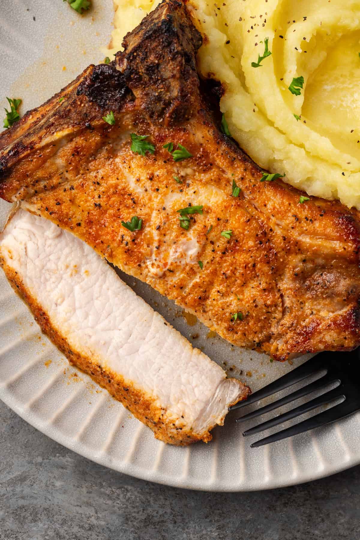 Make quick, easy, and juicy air fryer pork chops in 20 minutes! These bone-in chops are perfectly seared with a flavorful, caramelized crust. Close up of an air fryer pork chop with a slice cut from the end on a plate next to mashed potatoes.