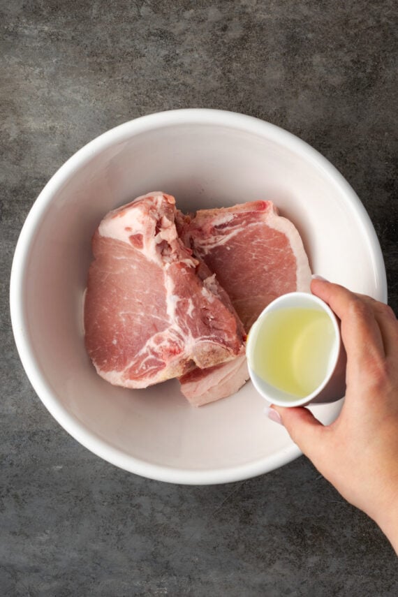 Make quick, easy, and juicy air fryer pork chops in 20 minutes! These bone-in chops are perfectly seared with a flavorful, caramelized crust. A hand pouring olive oil over two uncooked pork chops in a bowl.