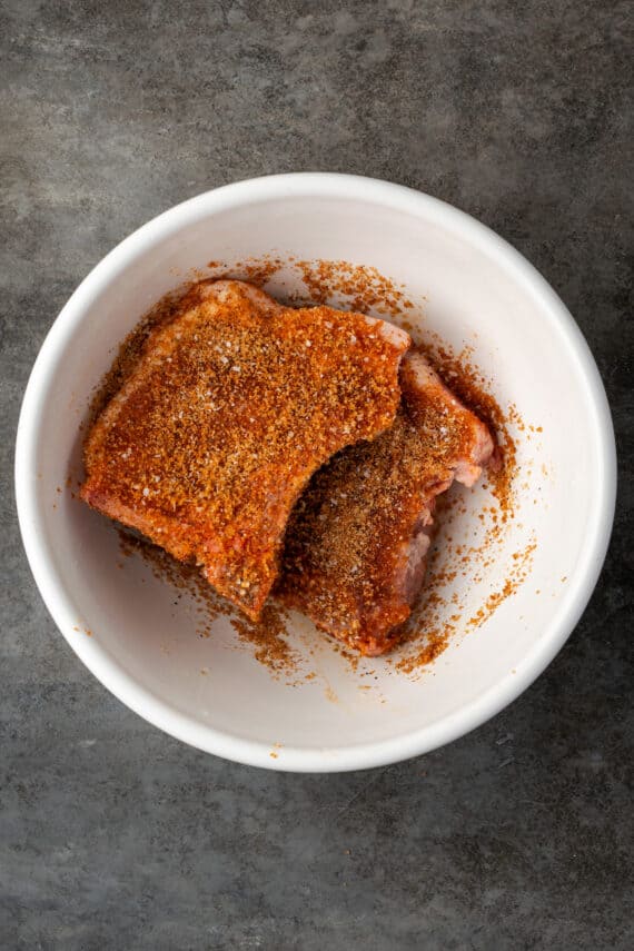 Make quick, easy, and juicy air fryer pork chops in 20 minutes! These bone-in chops are perfectly seared with a flavorful, caramelized crust. Two uncooked pork chops covered with seasoning in a bowl.