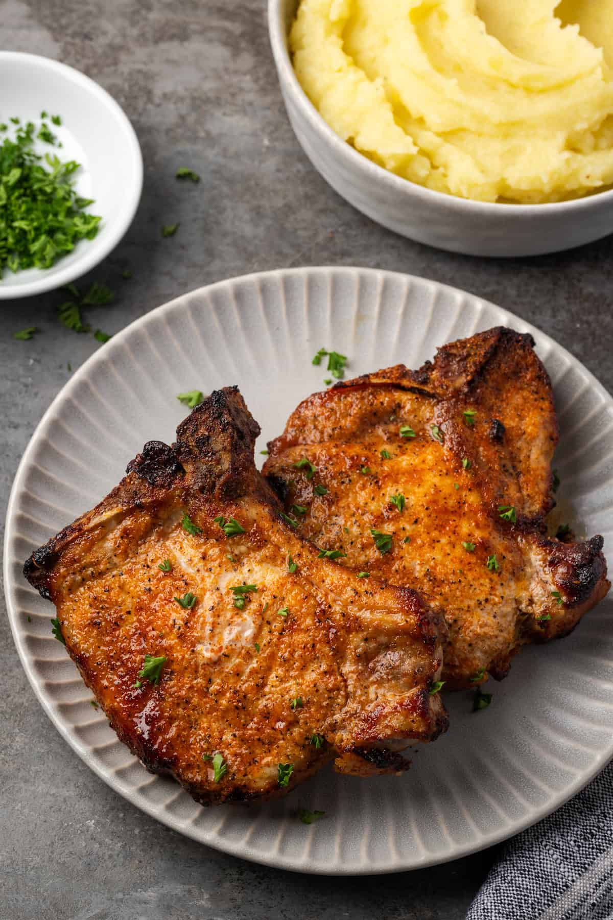 Make quick, easy, and juicy air fryer pork chops in 20 minutes! These bone-in chops are perfectly seared with a flavorful, caramelized crust. Overhead view of two air fryer pork chops on a plate, with a bowl of mashed potatoes in the background.