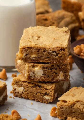 Three butterscotch blondies stacked next to a glass of milk.
