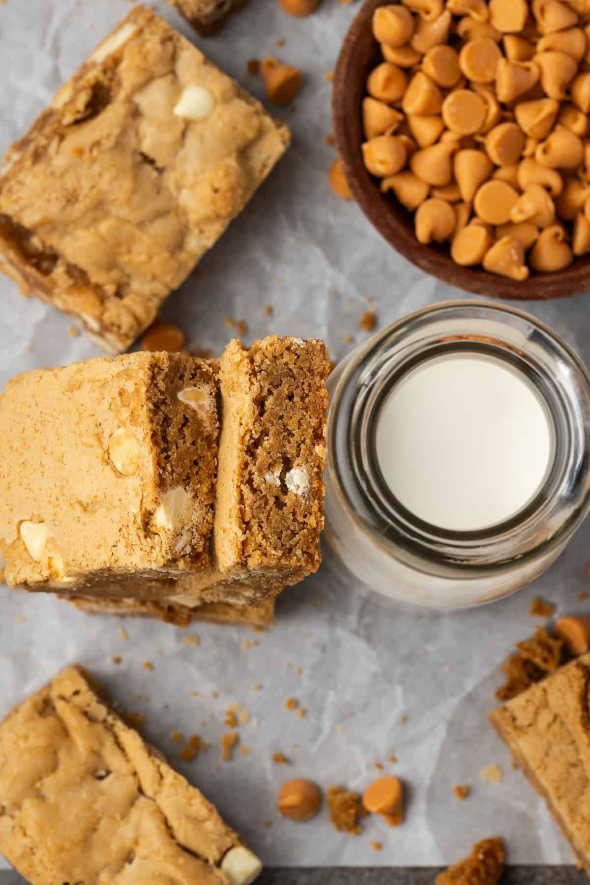 Overhead view of butterscotch blondies stacked next to a glass of milk and a bowl of butterscotch chips.