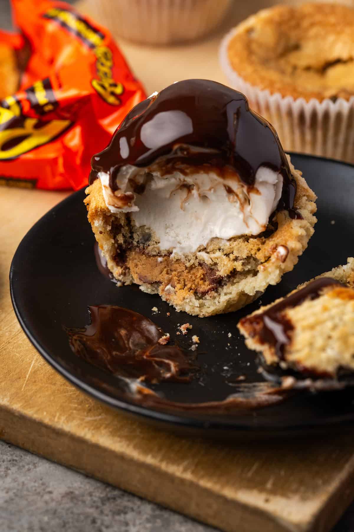 These are the ultimate cookie cups with two types of refrigerated cookie dough baked around a fudgy peanut butter cup. Quick and easy to make! A cookie cup topped with a scoop of ice cream and chocolate fudge sauce cut in half on a black ceramic plate.