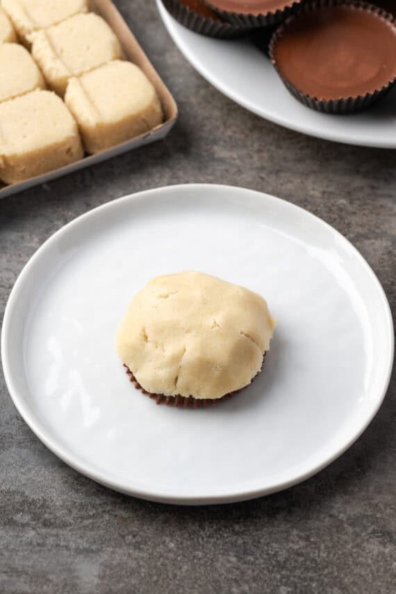 These are the ultimate cookie cups with two types of refrigerated cookie dough baked around a fudgy peanut butter cup. Quick and easy to make! A Reese's peanut butter cup face-down on a white plate, topped with sugar cookie dough.