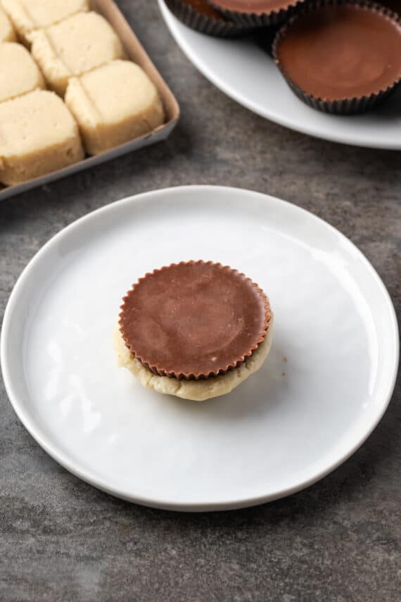 These are the ultimate cookie cups with two types of refrigerated cookie dough baked around a fudgy peanut butter cup. Quick and easy to make! A Reese's peanut butter cup with the bottom wrapped in sugar cookie dough on a plate.