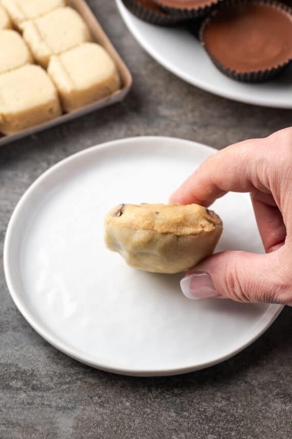 These are the ultimate cookie cups with two types of refrigerated cookie dough baked around a fudgy peanut butter cup. Quick and easy to make! A hand holding an unbaked cookie cup made from chocolate chip cookie dough and sugar cookie dough wrapped around a Reese's peanut butter cup, with a plate below.