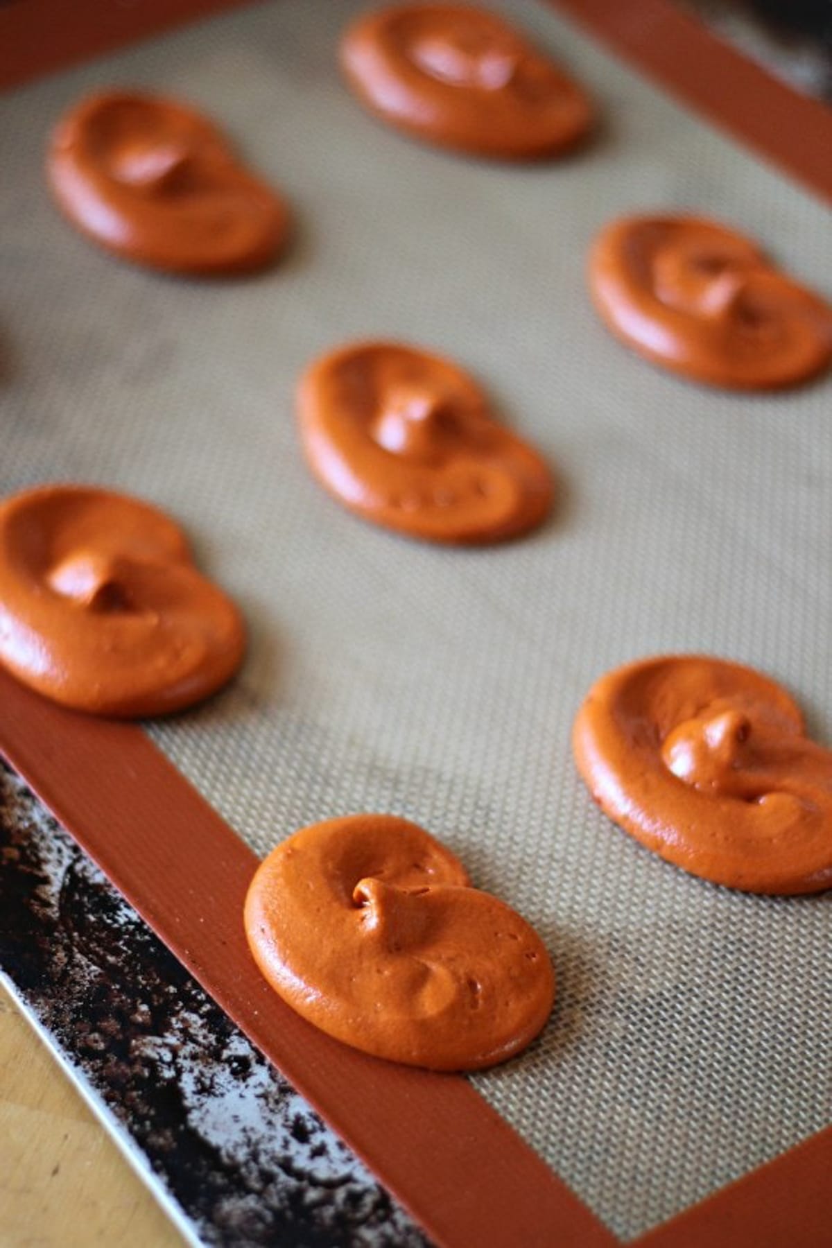 Orange batter piped on a silicone lined baking sheet in the shape of pumpkins