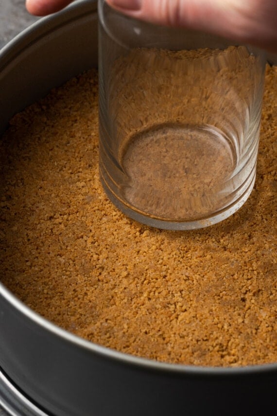 A hand uses the bottom of a glass to press a graham cracker crust into the bottom of a springform pan.