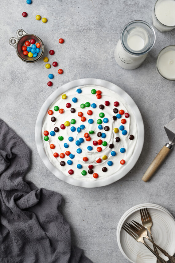 Top view of a whole candy pie topped with whipped cream and M&Ms, next to plates and forks.