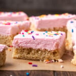 Sugar cookie bars topped with pink frosting and rainbow sprinkles on a parchment-lined cutting board.