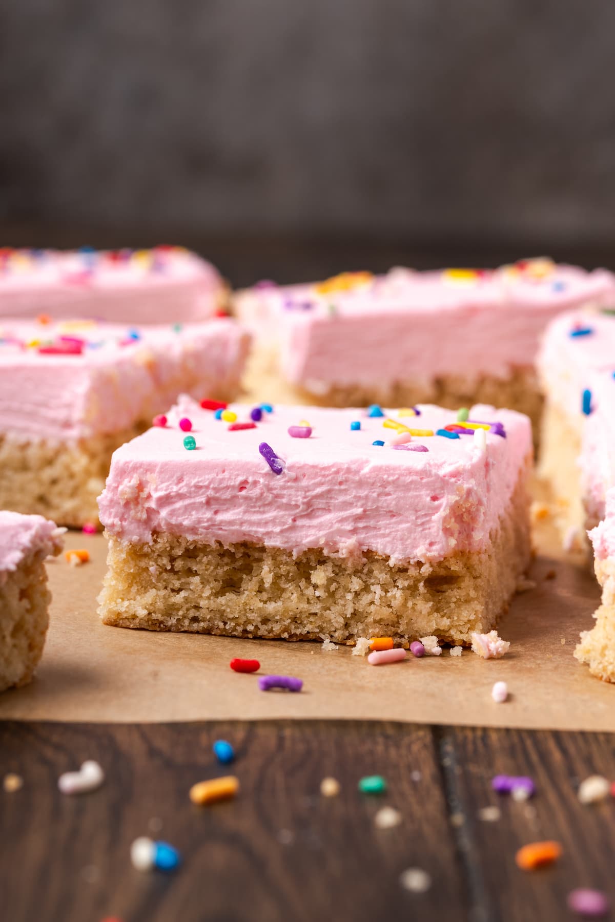 Sugar cookie bars topped with pink frosting and rainbow sprinkles on a parchment-lined cutting board.