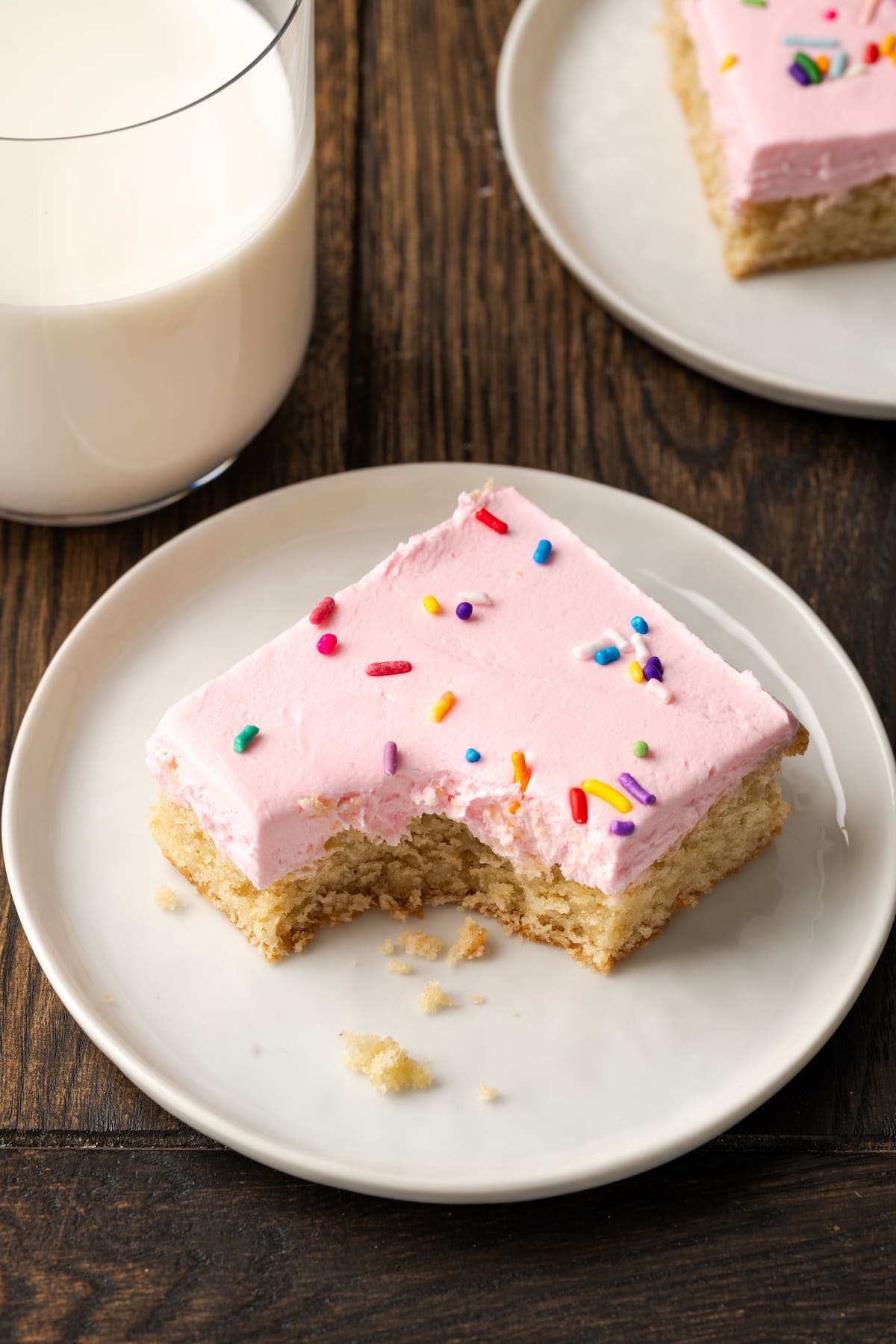A frosted sugar cookie bar with a bite missing on a white plate, next to a glass of milk.