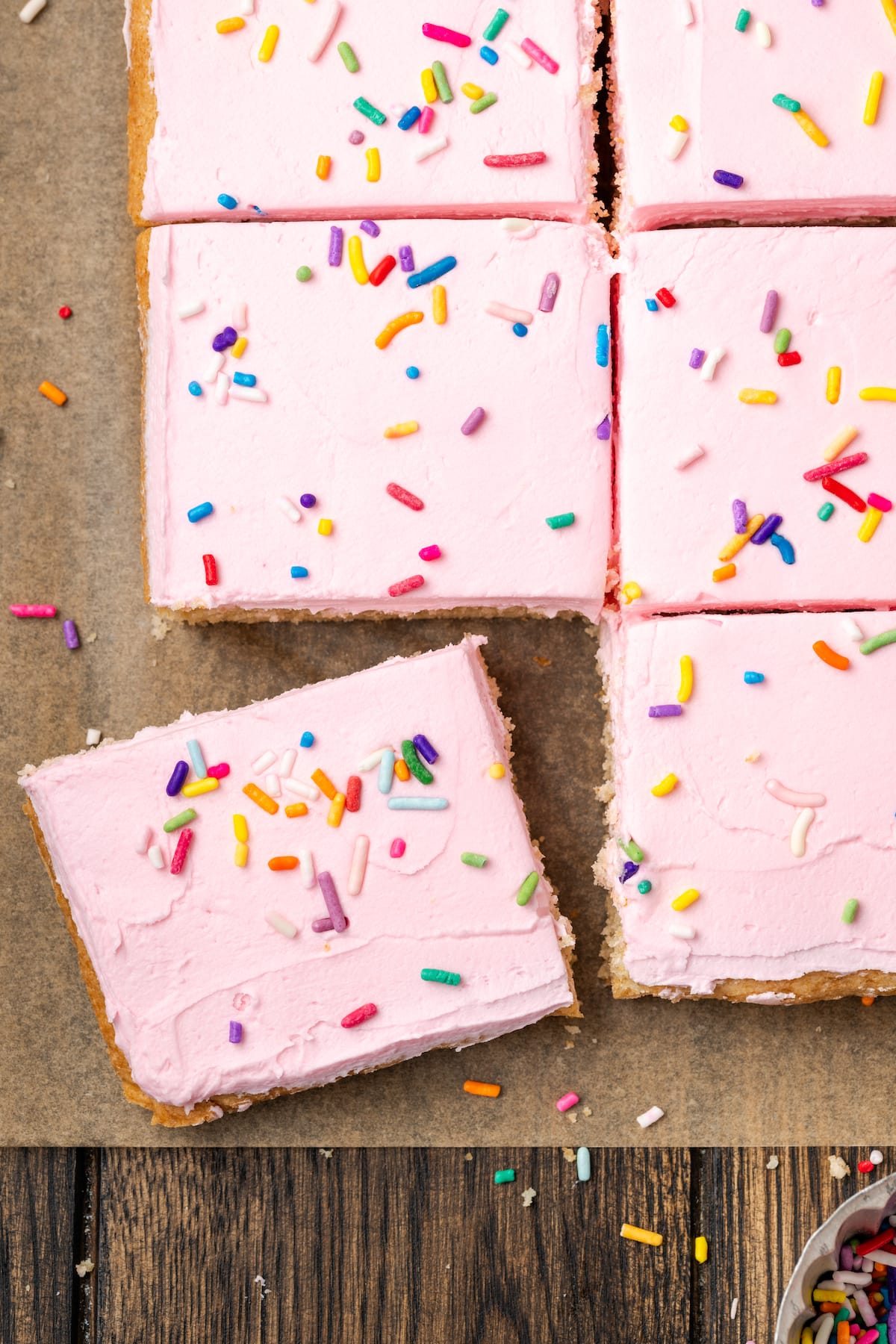 Overhead view of frosted sugar cookie bars topped with rainbow sprinkles cut into squares on a parchment-lined cutting board.