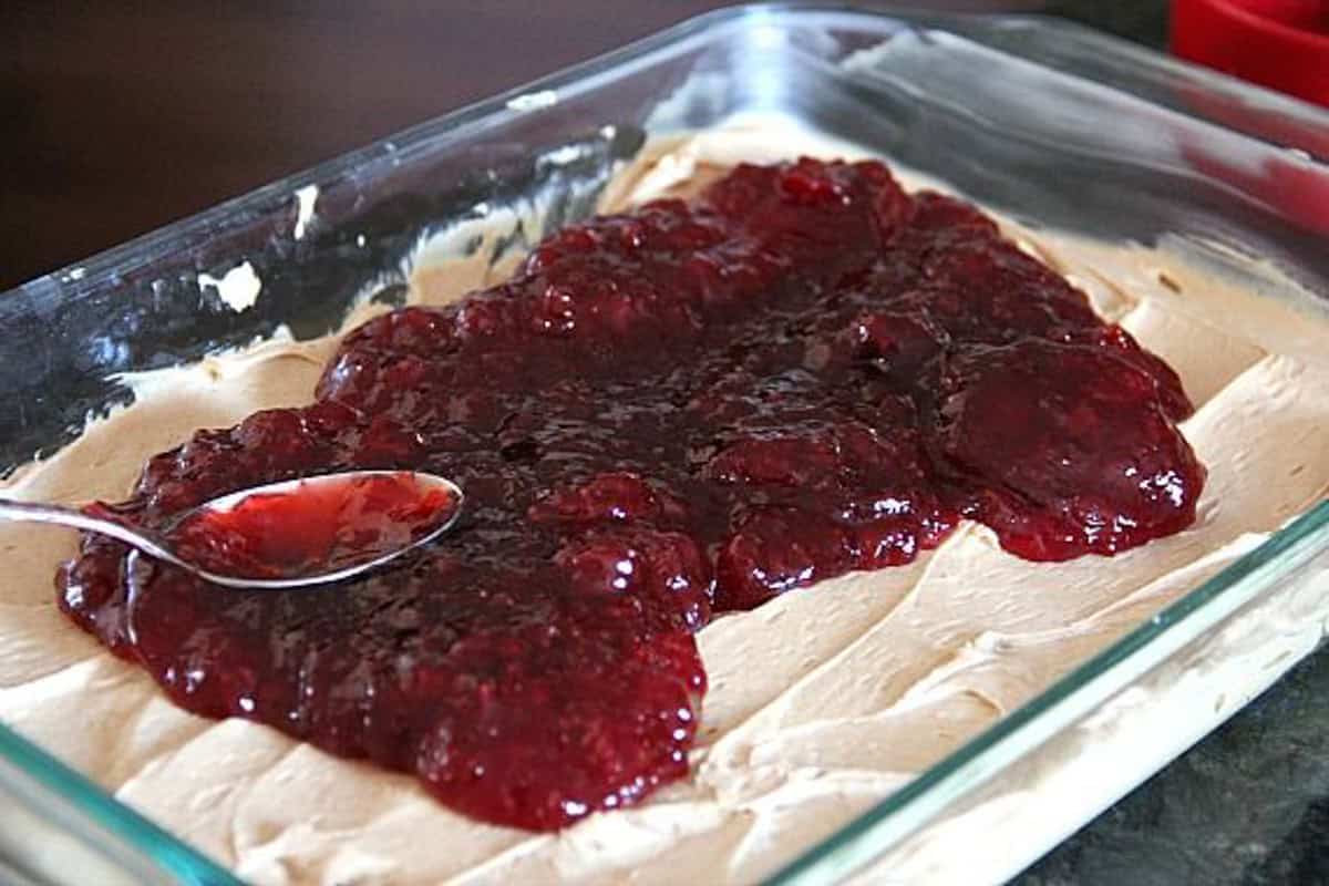 A spoon spreading jelly over the peanut butter layer in a glass baking dish.
