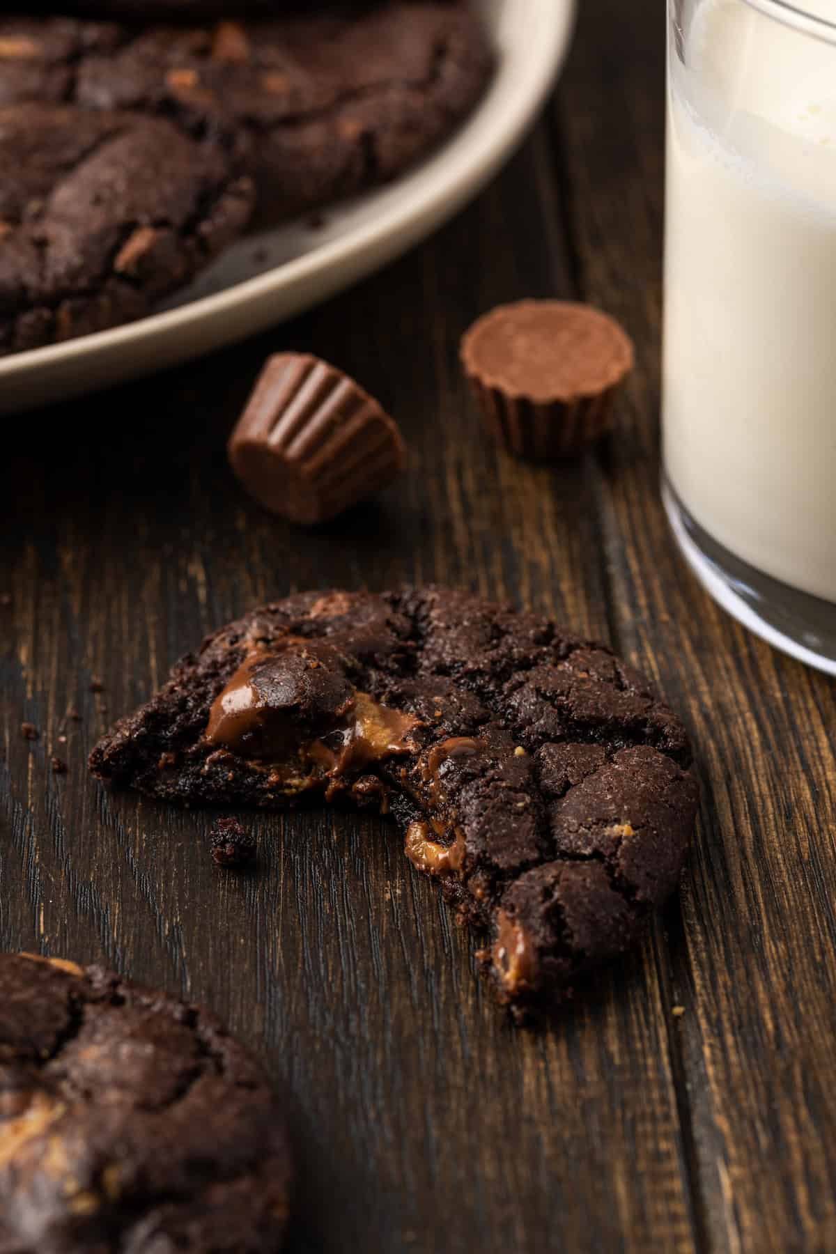 A peanut butter chocolate cookie with a bite missing on a wooden countertop, next to a plate of cookies and a glass of milk.