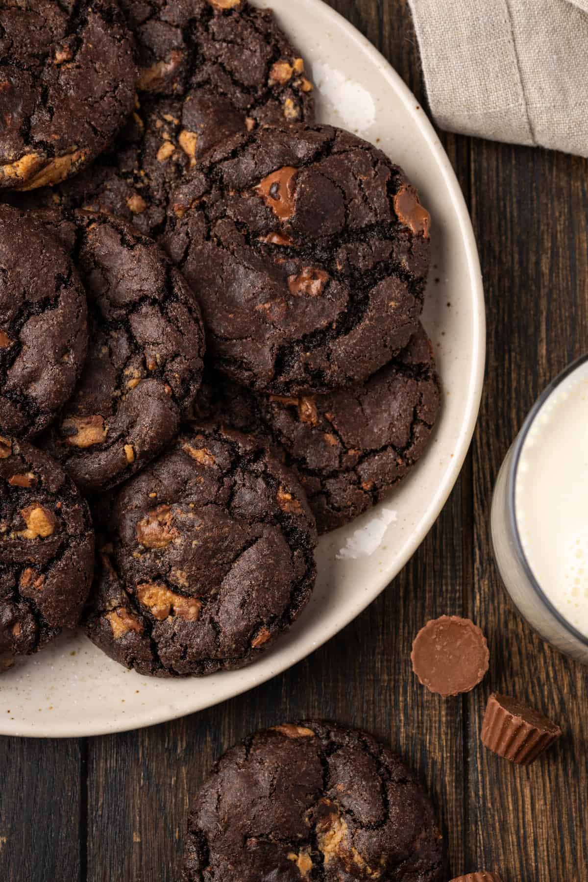 Overhead view of a pile of peanut butter chocolate cookies arranged on a white plate, next to a glass of milk.