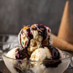 Three scoops of vanilla ice cream topped with chocolate sauce and sprinkles in a glass bowl, with ice cream cones in the background.