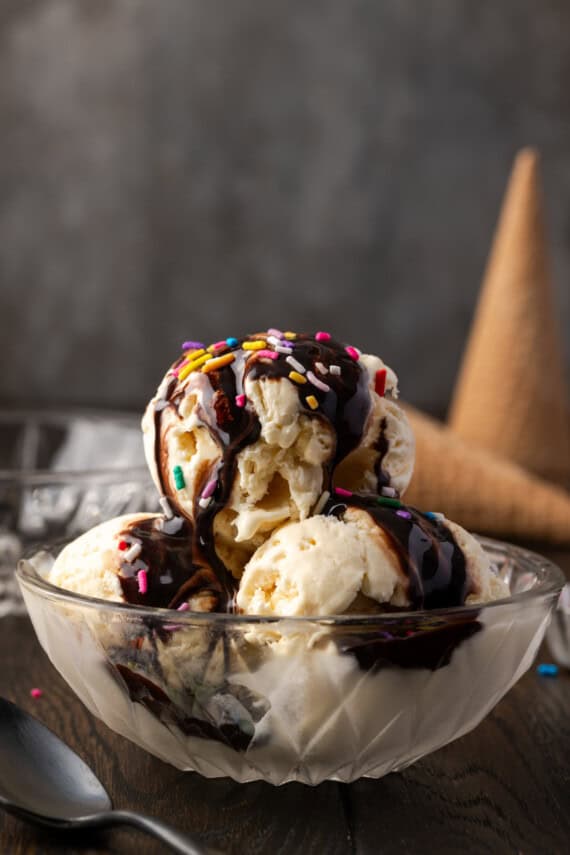 Three scoops of vanilla ice cream topped with chocolate sauce and sprinkles in a glass bowl, with ice cream cones in the background.