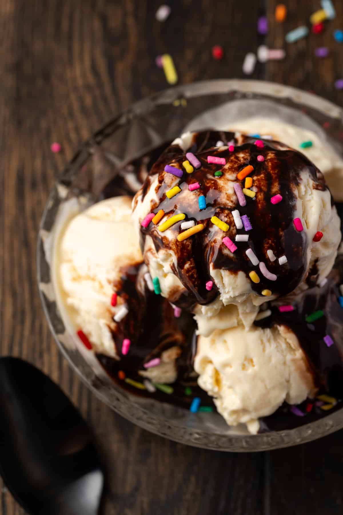 Close up overhead view of three scoops of vanilla ice cream topped with chocolate sauce and sprinkles in a glass bowl.