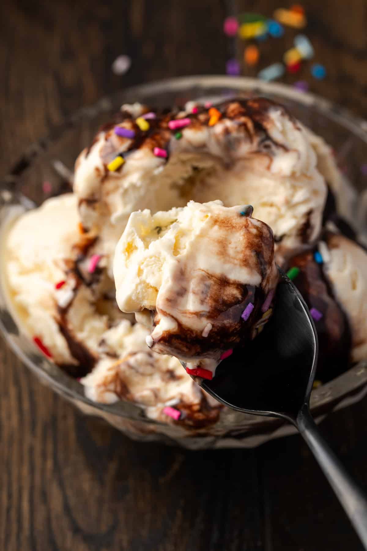 A spoonful of ice cream held over a bowl with scoops of ice cream covered with chocolate sauce and sprinkles.
