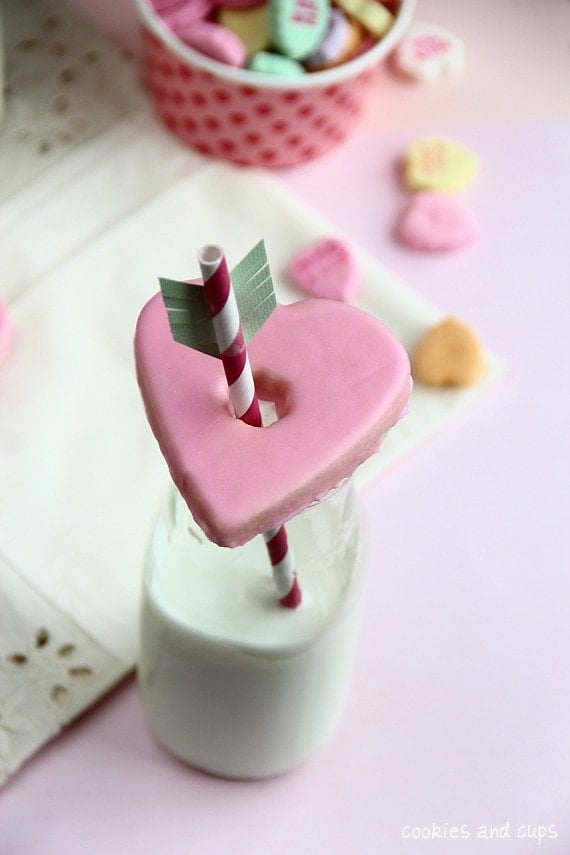 A heart-shaped Valentine's cookie on top of a glass of milk with a straw through the center.