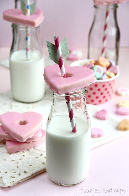 A heart-shaped Valentine's cookie on top of a glass of milk with a straw through the center.