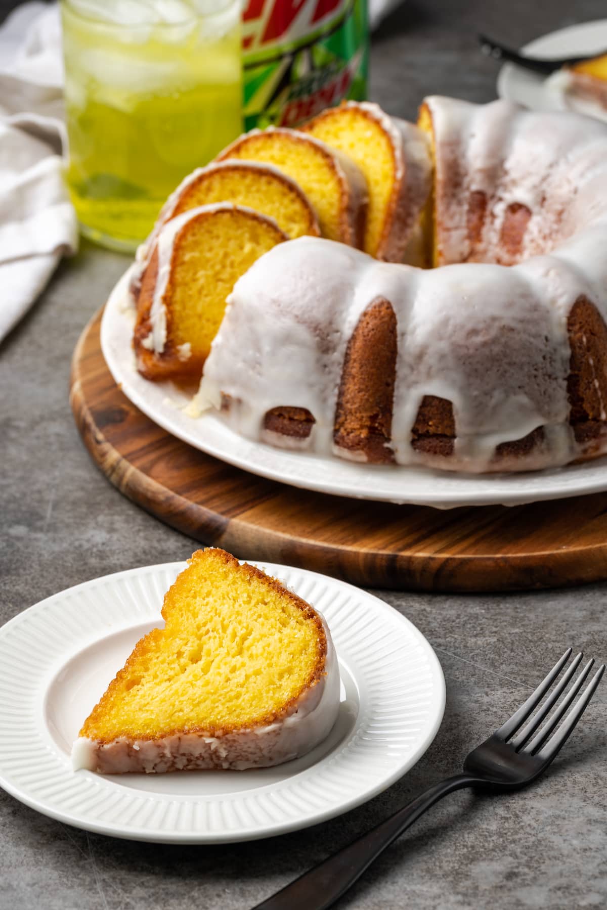 A slice of Mountain Dew cake on a white plate, with the rest of the bundt cake next to a glass of Mountain Dew in the background.