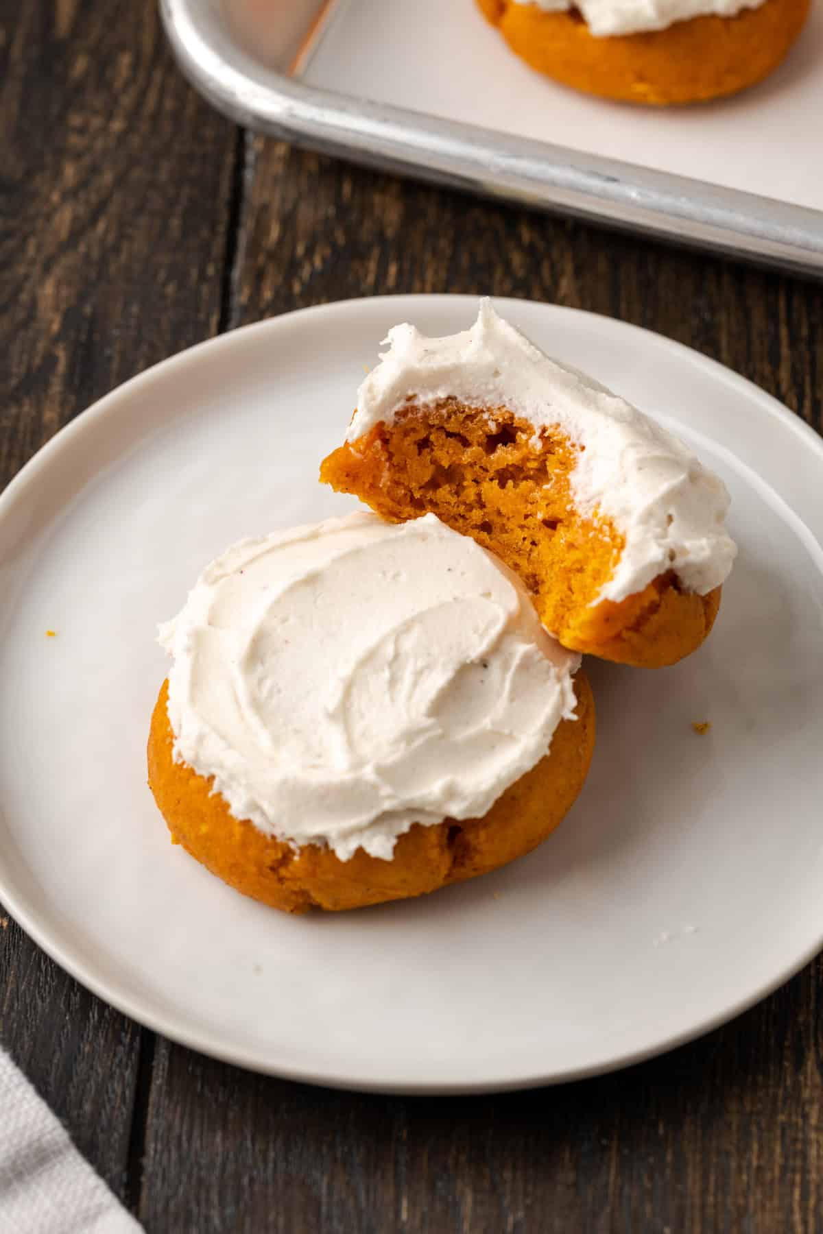 Two frosted cake mix pumpkin cookies on a white plate, with a bite missing from one cookie.