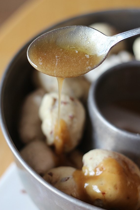 A mixture of melted butter and brown sugar drips from a spoon, with a bundt pan filled with cinnamon roll monkey bread in the background