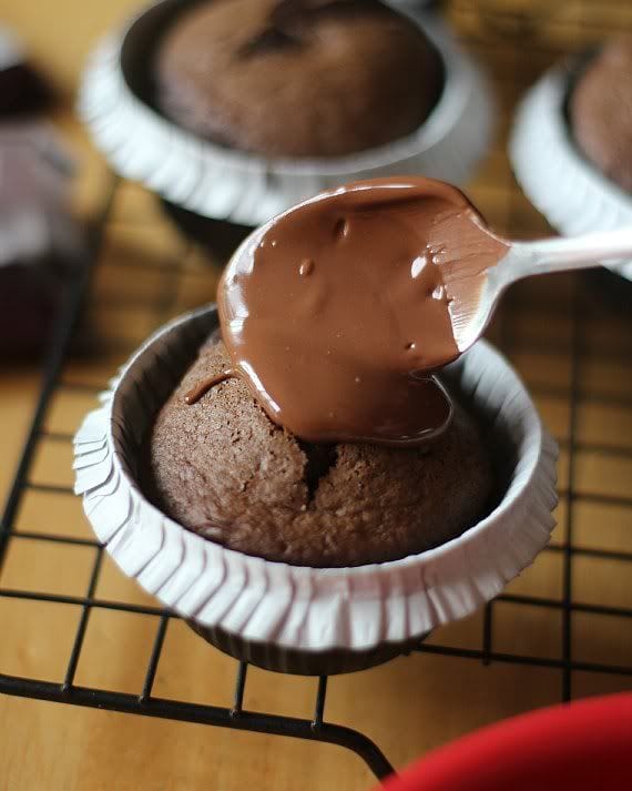 Melted chocolate being spooned over a chocolate cupcake on a cooling rack