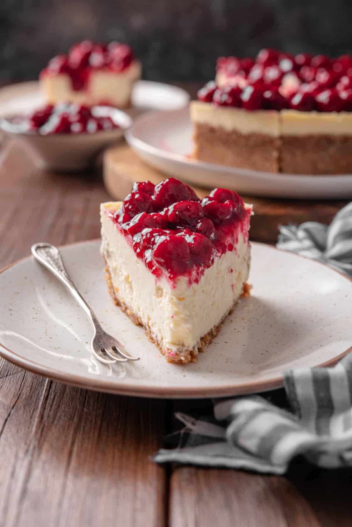 A fork resting next to a slice of cherry cheesecake on a plate, with the rest of the cheesecake in the background.