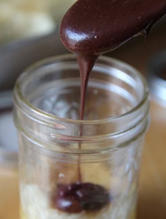 A spoonful of chocolate sauce being layered in a jar with vanilla pudding and vanilla cake