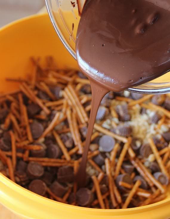 Melted chocolate being poured over a bowl of pretzel sticks, Rolo candies, Mini Peanut Butter Cups and Crispy Rice cereal