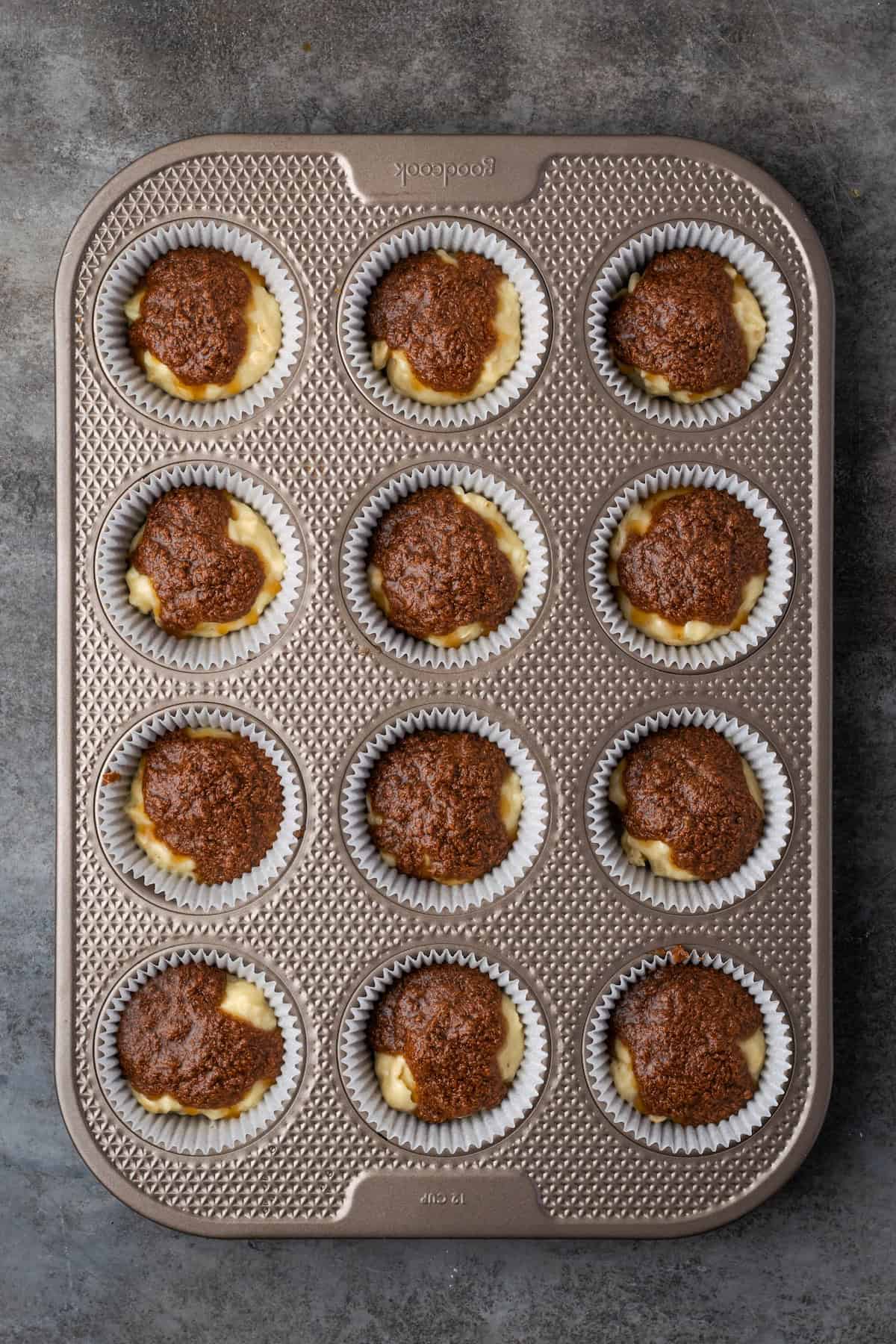 Cinnamon muffin batter topped with cinnamon sugar filling in a 12-well muffin pan.