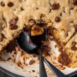 Overhead view of a skillet chocolate chip cookie with a serving missing and a spoon resting in the open space.