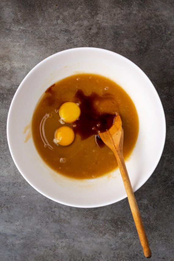 Egg and vanilla added to wet cookie dough ingredients in a white bowl with a wooden spoon.