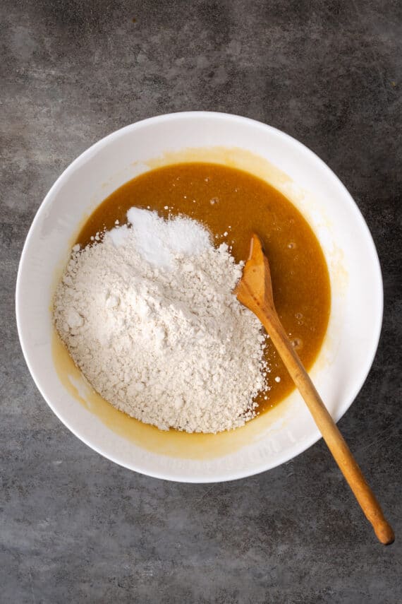 Dry ingredients added to wet cookie dough ingredients in a white bowl with a wooden spoon.