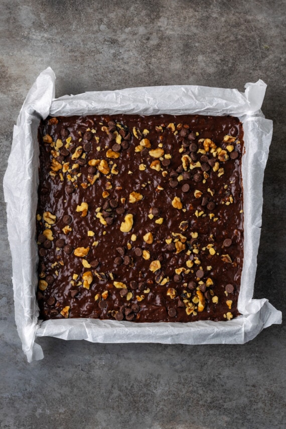 Banana bread brownie batter topped with chopped nuts in a square baking pan lined with parchment paper.