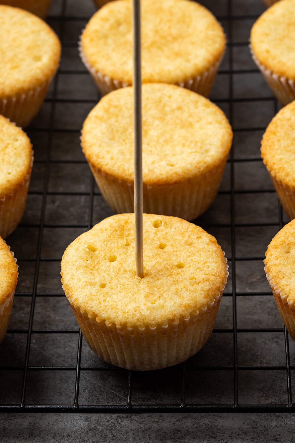 A skewer poking holes in the top of a baked vanilla cupcake.
