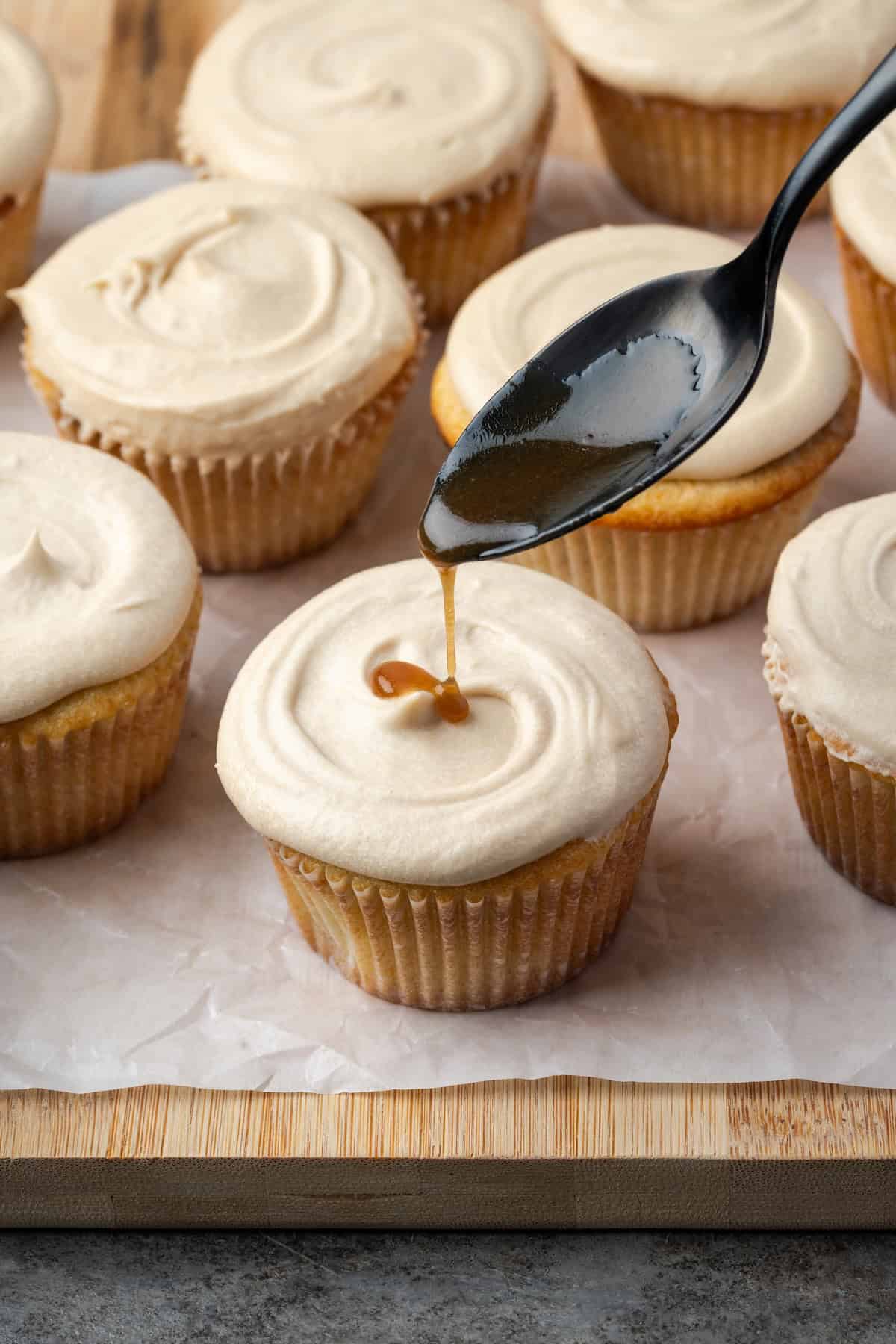 A spoon drizzling caramel sauce over a frosted salted caramel cupcake, surrounded by more cupcakes on a parchment-lined wooden cutting board.