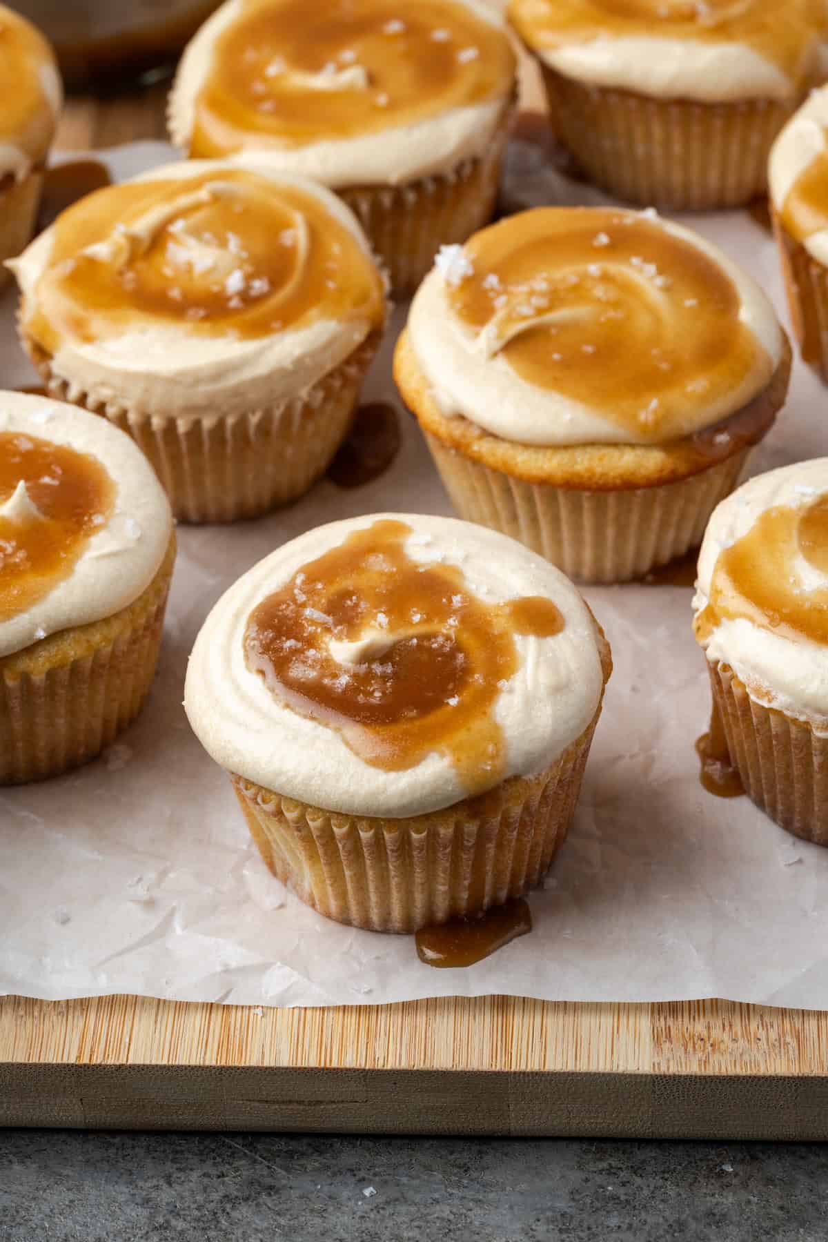Frosted salted caramel cupcakes topped with caramel and sea salt on a parchment-lined wooden cutting board.
