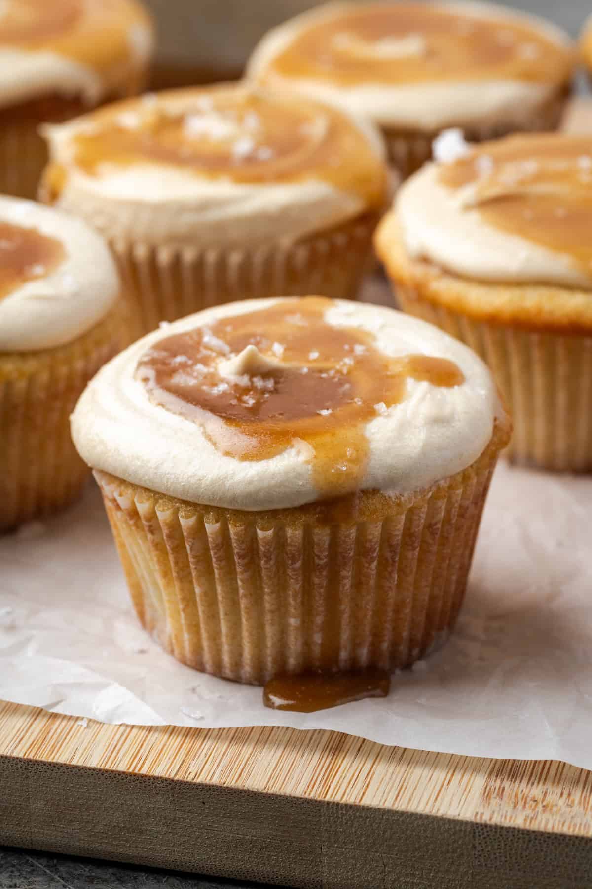 These fluffy homemade salted caramel cupcakes are soaked in caramel sauce, frosted with caramel buttercream, and finished with sea salt. Salted caramel cupcakes on a parchment-lined wooden cutting board.