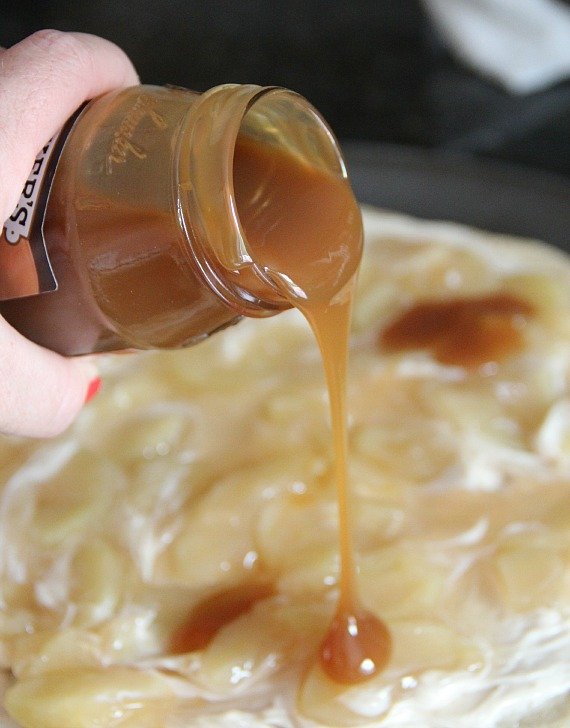 Caramel sauce being poured over giant sugar cookie with cream cheese and apple topping