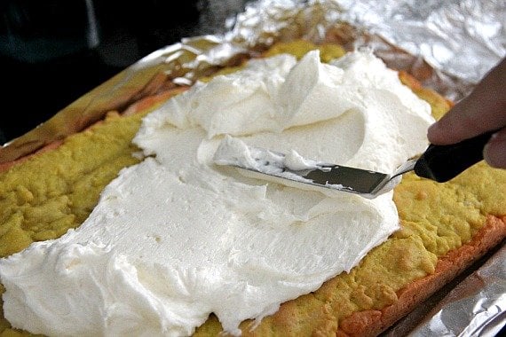 White frosting being spread over yellow and orange layered bars in a baking pan