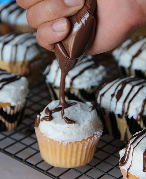 Melted chocolate being piped on top of vanilla frosted cupcakes