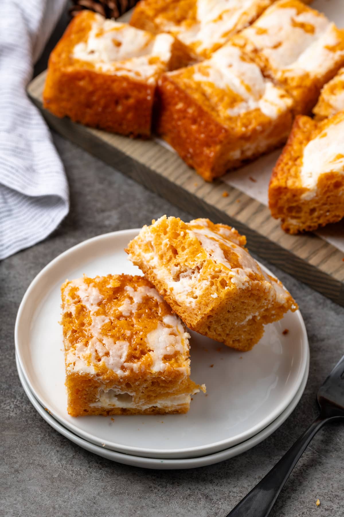 Two pumpkin cream cheese bars on a white plate next to more bars on a wooden board.