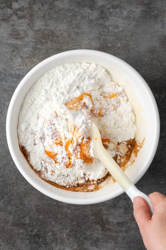 A hand uses a spatula to stir together pumpkin batter in a metal mixing bowl.